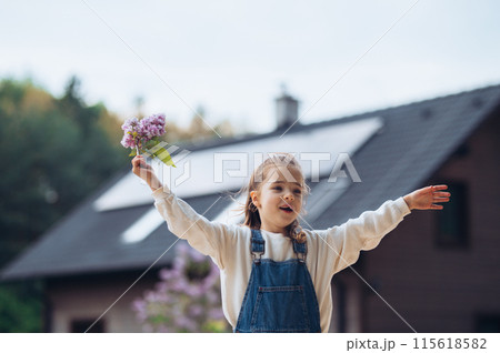 Beautiful young girl with purple lilacs flower in hands. Young girl spending time outdoors in nature, springtime. House with solar panels in background. Beautiful young girl with purple lilacs flower in hands. Young girl spending time outdoors in nature, springtime. House with solar panels in background. 115618582
