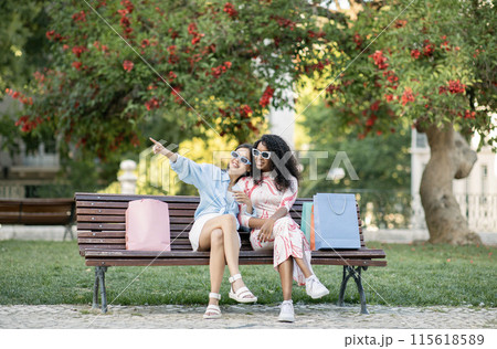 Two Happy Multiethnic Female Friends Relaxing On Bench After Shopping, Cheerful Young Women In Stylish Eyeglasses Resting Outdoors With Purchases After Walk In Summer City, Enjoying Seasonal Sales 115618589