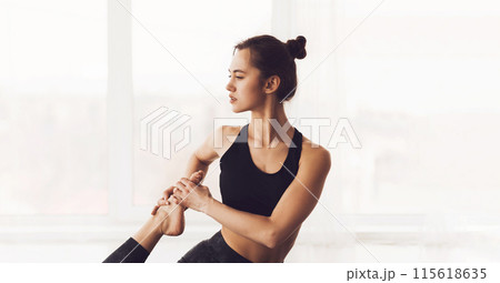 A focused young woman is seated on the floor in a well-lit room, extending one leg while holding her foot with both hands as part of a stretching routine. A focused young woman is seated on the floor in a well-lit room, extending one leg while holding her foot with both hands as part of a stretching routine. 115618635