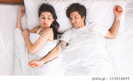 A young couple is sleeping together in a white bed. The woman is on her side, facing the man, with her arm wrapped around her body. 115618645