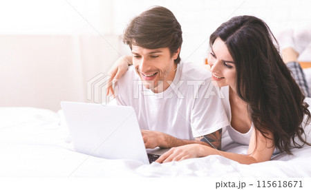 A young couple is lying in bed, using a laptop computer. The man is looking at the screen with a smile on his face while the woman is looking at him, also smiling A young couple is lying in bed, using a laptop computer. The man is looking at the screen with a smile on his face while the woman is looking at him, also smiling 115618671
