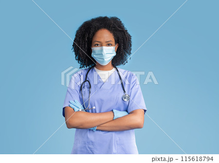 Serious confident young black woman doctor, nurse in blue uniform , protective mask and gloves, crossed hands on chest, isolated on blue studio background. Medicine, health care, treatment 115618794