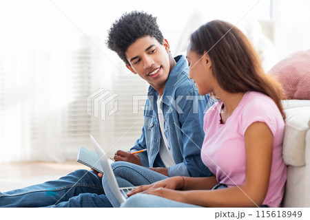 African American teen couple sits on the floor, the guy is working on a laptop while the girl is writing in a notepad. They are engaged in a conversation, the guy seems to be explaining something African American teen couple sits on the floor, the guy is working on a laptop while the girl is writing in a notepad. They are engaged in a conversation, the guy seems to be explaining something 115618949
