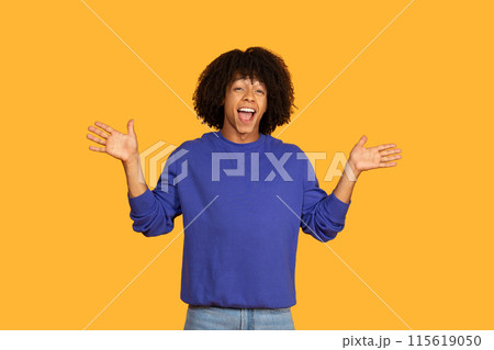 A young guy with curly hair is exuberantly expressing joy with an energetic pose. They are dressed in a blue sweater and jeans, standing in front of a bold yellow backdrop. A young guy with curly hair is exuberantly expressing joy with an energetic pose. They are dressed in a blue sweater and jeans, standing in front of a bold yellow backdrop. 115619050