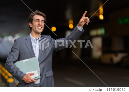 Happy handsome young businessman wearing formal outfit raising han up, catching taxi on the street, holding laptop and smartphone, entrepreneur going to business meeting, copy space 115619082