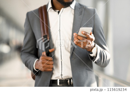 An African American man is using his phone in an airport terminal, standing with his luggage. The setting is modern and bustling, indicating efficient travel, cropped An African American man is using his phone in an airport terminal, standing with his luggage. The setting is modern and bustling, indicating efficient travel, cropped 115619234