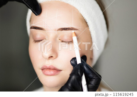 Professional Cosmetologist Mapping Eyebrows Of Young Woman With White Pencil Before Permanent Makeup Treatment In Beauty Salon, Marking Up Brow Zone And Forehead Before PMU Procedure, Closeup Shot 115619245