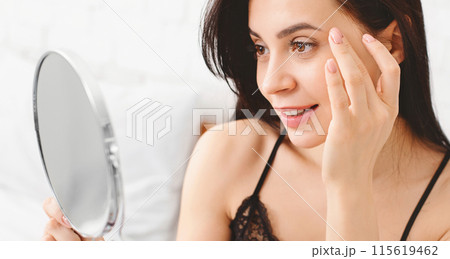 Young millennial woman using mirror and touching her face in bedroom in morning, free space 115619462