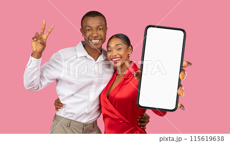 A happy, young black couple stands in front of a pink background. The man making a peace sign with his right hand. The woman holding a smartphone with a blank white screen 115619638