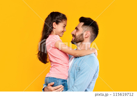 Father's love. Loving father holding his cute little daughter, dad bonding with her child girl, having fun with kid over yellow background, side view, copy space Father's love. Loving father holding his cute little daughter, dad bonding with her child girl, having fun with kid over yellow background, side view, copy space 115619712