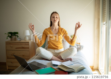 A teen girl is sitting on her bed, meditating with closed eyes and a serene expression. She is surrounded by open books, a laptop, and notebooks, indicating she is taking a break from studying A teen girl is sitting on her bed, meditating with closed eyes and a serene expression. She is surrounded by open books, a laptop, and notebooks, indicating she is taking a break from studying 115619756