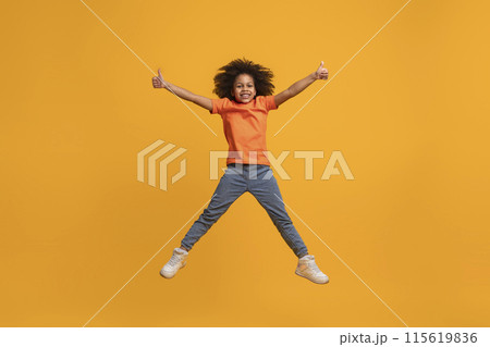 African American girl jumping in the air with her arms outstretched and giving a thumbs up with both hands. She is smiling and appears happy. The background is a solid yellow color. 115619836