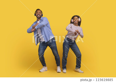 African American man and woman are captured mid-dance, both smiling and enjoying themselves as they perform a coordinated movement, using headphones on yellow background African American man and woman are captured mid-dance, both smiling and enjoying themselves as they perform a coordinated movement, using headphones on yellow background 115619885