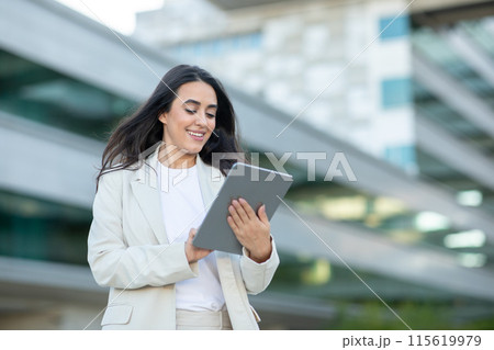 A businesswoman in a white blazer stands in front of a modern office building while using her tablet. She smiles as she looks at the screen, demonstrating her confident and successful demeanor 115619979