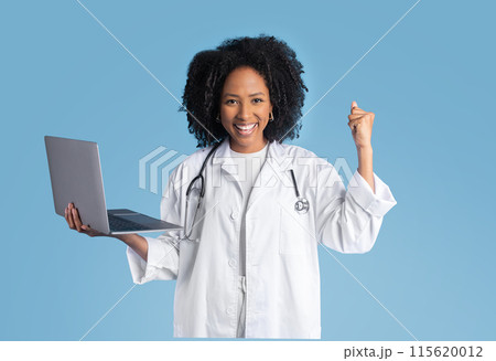 Satisfied young black lady doctor therapist in white coat hold laptop, making success hand gesture, celebrating victory, isolated on blue studio background. Medical good result, health care 115620012