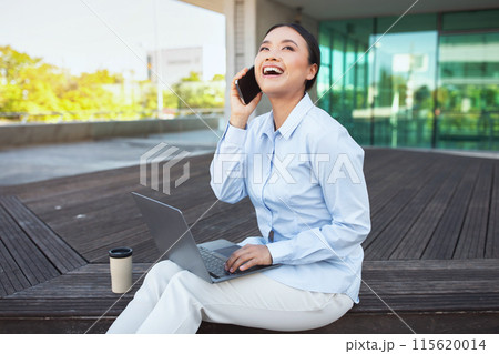 Asian woman is seated on a bench, engaged in conversation on her phone, holding computer on her lap. She appears focused, possibly discussing important matters 115620014