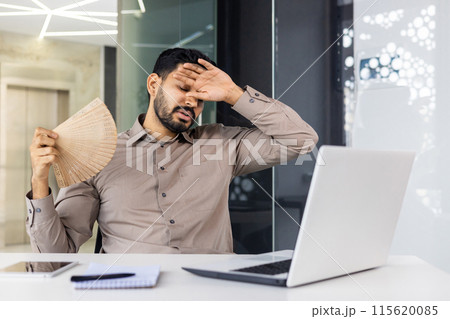 Businessman in brown shirt fanning himself, feeling stressed due to heat in a modern office setting while working on a laptop. 115620085