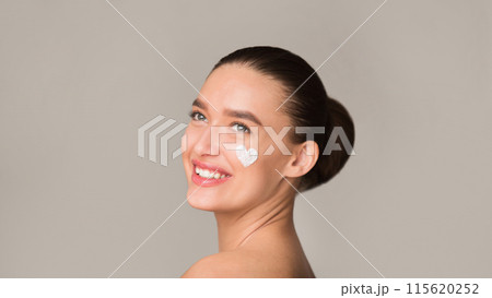 A woman is smiling while wearing a cream mask on her face. The cream is applied evenly, covering her skin from forehead to chin. Her smile is bright and genuine, coseup 115620252