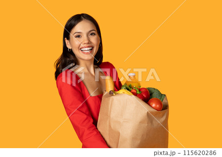 Happy young woman is standing outdoors, holding a paper bag filled with groceries. She appears to be carrying the bag effortlessly, showcasing a sense of independence and responsibility. Happy young woman is standing outdoors, holding a paper bag filled with groceries. She appears to be carrying the bag effortlessly, showcasing a sense of independence and responsibility. 115620286