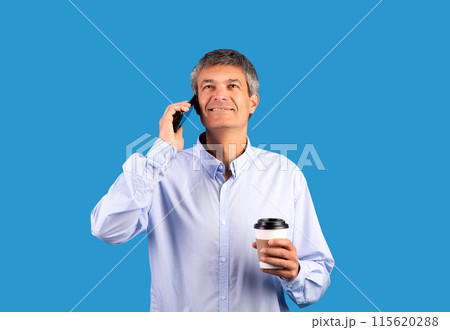 Smiling gray haired man talking on cellphone, communicating and holding a coffee cup standing on blue studio background. Mature businessman having pleasant conversation and enjoying hot drink 115620288