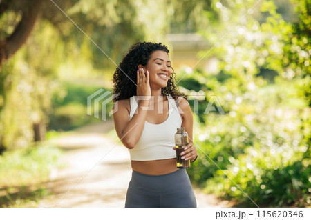 A woman with curly hair stands in a sunlit park, smiling and holding a water bottle. She appears to be on a morning workout, enjoying the fresh air and nature around her. A woman with curly hair stands in a sunlit park, smiling and holding a water bottle. She appears to be on a morning workout, enjoying the fresh air and nature around her. 115620346