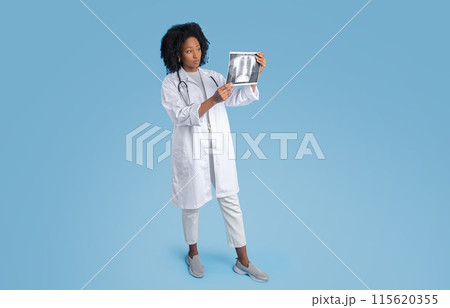 Serious young black woman doctor in white coat looking at fluorography shot, isolated on blue studio background. Medical exam, health care, diagnostic and ill help, full length Serious young black woman doctor in white coat looking at fluorography shot, isolated on blue studio background. Medical exam, health care, diagnostic and ill help, full length 115620355