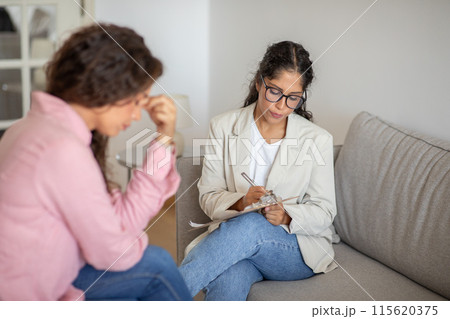 A woman in a white blazer and blue jeans sits on a couch and takes notes with a pen and clipboard. She is listening intently to the woman across from her, who is sitting with her head down A woman in a white blazer and blue jeans sits on a couch and takes notes with a pen and clipboard. She is listening intently to the woman across from her, who is sitting with her head down 115620375