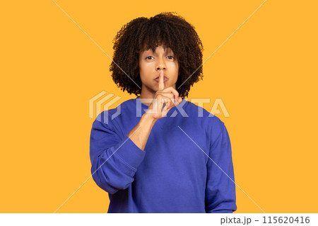A man with curly black hair is wearing a blue sweatshirt and gesturing for silence with his finger to his lips against a yellow background. A man with curly black hair is wearing a blue sweatshirt and gesturing for silence with his finger to his lips against a yellow background. 115620416