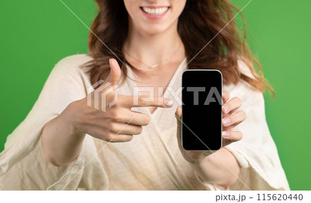Happy young european lady point finger at smartphone with blank screen, isolated on green studio background, close up. Recommendation sale, website and app for shopping, ad and offer 115620440