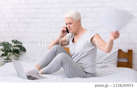 Upset senior woman talking on phone and working at home, looking at laptop and paper correspondence, sitting on bed at home, panorama Upset senior woman talking on phone and working at home, looking at laptop and paper correspondence, sitting on bed at home, panorama 115620455