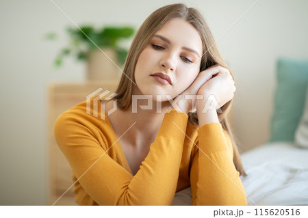 A teen girl, dressed in a yellow shirt, sits indoors with a thoughtful expression on her face. The background is softly lit, and a potted plant is visible behind her 115620516