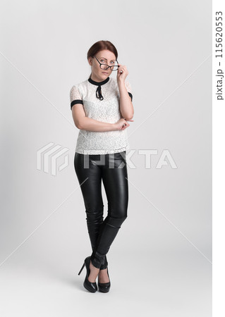 Businesswoman looking at camera over eyeglasses, which she holds with one hand near face. Business person wearing blouse, trousers and high heels standing legs crossed at ankle on white background Businesswoman looking at camera over eyeglasses, which she holds with one hand near face. Business person wearing blouse, trousers and high heels standing legs crossed at ankle on white background 115620553