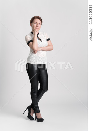 Business woman wearing white short sleeve blouse, black leather tight trousers and high heeled shoes looking at camera with one hand raised to chin, standing legs crossed at ankle on white background 115620555