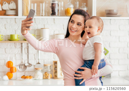 A woman holding her baby in her arms while taking a selfie with a smartphone. The mother and child are smiling at the camera in a casual and joyful moment. 115620560
