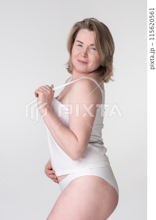 Serious woman in white underwear puts hand on stomach while holding strap of camisole. Overweight middle aged adult woman looking over shoulder at camera, represents women's healthy lifestyle concept 115620561