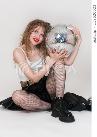 Cheerful young adult pin up girl holding and presses mirror disco ball to herself, looking at camera. Smiling woman dressed in crop top, shorts and boots in clubbing style, sitting on white background Cheerful young adult pin up girl holding and presses mirror disco ball to herself, looking at camera. Smiling woman dressed in crop top, shorts and boots in clubbing style, sitting on white background 115620623