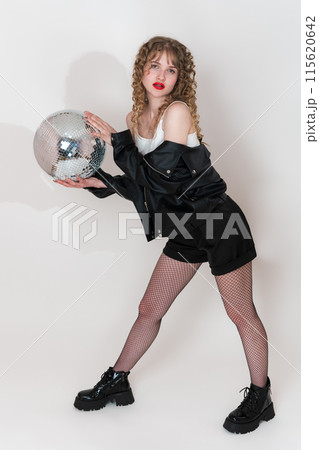 Confident woman holding disco mirror ball and looking at camera, standing on white. Beautiful young adult pin up girl with long curly hair dressed in black jacket, shorts and boots in clubbing style 115620642