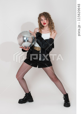 Portrait of serious young woman dancer holding mirror disco ball, looking at camera. Young adult pin up girl with long curly hair dressed clubbing in black leather jacket, shorts, crop top and boots Portrait of serious young woman dancer holding mirror disco ball, looking at camera. Young adult pin up girl with long curly hair dressed clubbing in black leather jacket, shorts, crop top and boots 115620648