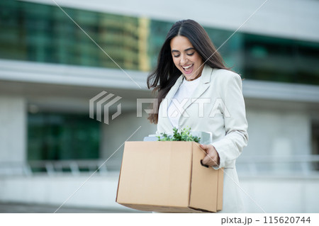 A young businesswoman, dressed in a white blazer and white shirt, walks away from her office building while carrying a cardboard box filled with her belongings 115620744