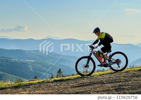 Cyclist man riding electric mountain bike outdoors. Male tourist biking along grassy trail in the mountains, wearing helmet and backpack. Concept of sport, active leisure and nature. Cyclist man riding electric mountain bike outdoors. Male tourist biking along grassy trail in the mountains, wearing helmet and backpack. Concept of sport, active leisure and nature. 115620756