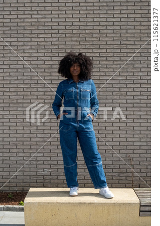 Confident woman in denim jumpsuit, against brick wall, showcasing stylish fashion sense Confident woman in denim jumpsuit, against brick wall, showcasing stylish fashion sense 115621377