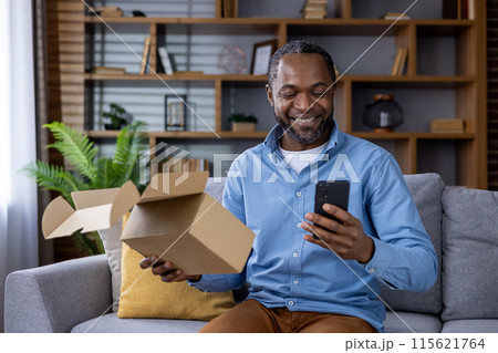 Smiling man unboxing a package and using a smartphone while relaxing on the sofa at home, enjoying a delivery. Smiling man unboxing a package and using a smartphone while relaxing on the sofa at home, enjoying a delivery. 115621764