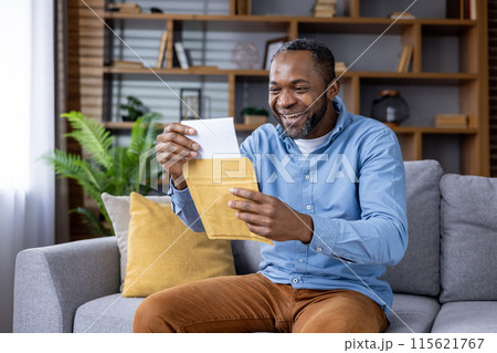 Joyful man wearing casual clothes and smiling while opening a letter on a sofa in a cozy living room. Joyful man wearing casual clothes and smiling while opening a letter on a sofa in a cozy living room. 115621767