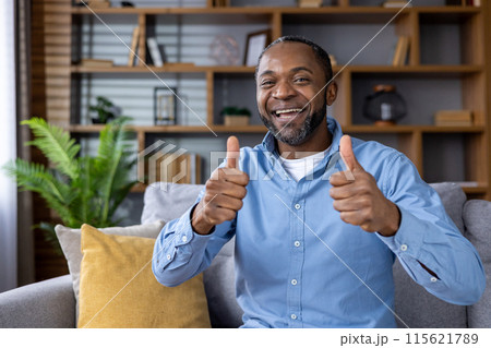 Cheerful man smiling and giving a thumbs up gesture while sitting on a sofa in a comfortable home environment ,with shelves and plants in the background. 115621789