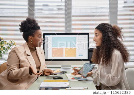Side view of two businesswomen discussing new mobile app interface design while holding color swatches. Diverse team talk shop against window in office, copy space 115622389