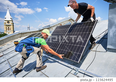 Workers building solar panel system on metal rooftop in urban setting. Two men installers carrying photovoltaic solar module outdoors. Alternative, green and renewable energy generation concept. 115624495