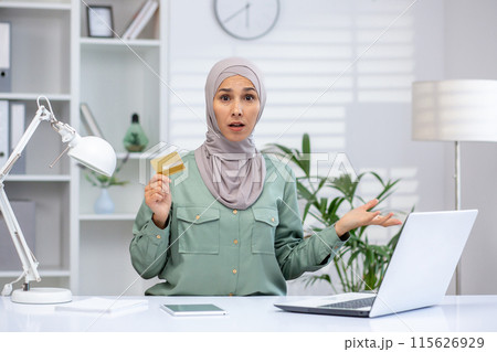 Confused woman in hijab holding credit card while using laptop in modern office setting. 115626929