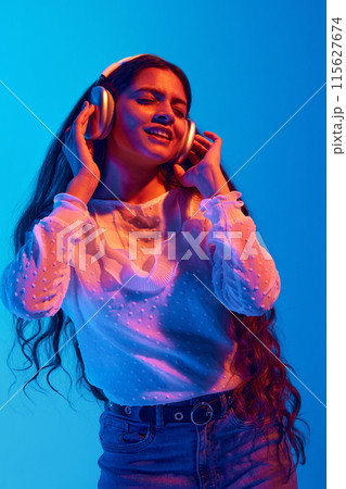 Creative portrait of young Indian woman in headphones dancing and singing while listening music in neon light against blue gradient background. Creative portrait of young Indian woman in headphones dancing and singing while listening music in neon light against blue gradient background. 115627674