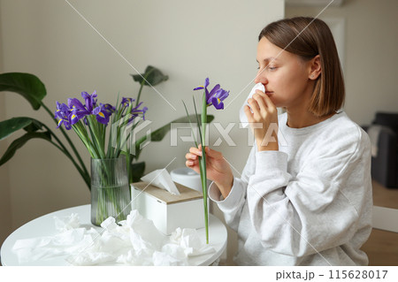 Allergic young woman holds iris flower, covers nose with paper tissue has runny nose, sneezes from flowers pollen at kitchen home by table. Girl with flu, itching or cough, seasonal allergy, rhinitis Allergic young woman holds iris flower, covers nose with paper tissue has runny nose, sneezes from flowers pollen at kitchen home by table. Girl with flu, itching or cough, seasonal allergy, rhinitis 115628017