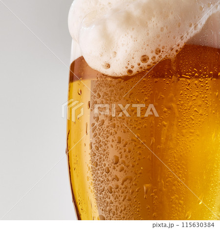 Close-up of pint glass filled with golden, amber beer topped with frothy white foam against grey studio background. Close-up of pint glass filled with golden, amber beer topped with frothy white foam against grey studio background. 115630384
